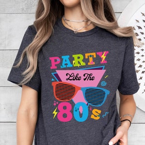 May include: A dark grey t-shirt with a colourful graphic design that says "Party Like The 80's". The graphic features a pair of sunglasses with a lightning bolt and a musical note.