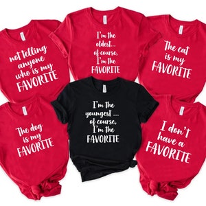 May include: Six red t-shirts with white text. The shirts have different sayings on them, including "I'm the oldest... of course, I'm the favorite", "The cat is my favorite", "I don't have a favorite", "The dog is my favorite", "I'm the youngest... of course, I'm the favorite", and "Not telling anyone who is my favorite".