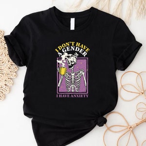 May include: Black t-shirt with a skeleton holding a cup of coffee. The text on the shirt reads "I don't have a gender I have anxiety".