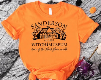 May include: An orange t-shirt with a black graphic featuring the text "Sanderson Witch Museum" and an illustration of a house. The text "home of the black flame candle" is also included. The shirt is styled for Halloween.