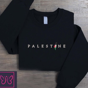 May include: Black crew neck sweatshirt with the word "PALESTINE" in white capital letters. The letter "E" has a small graphic of the Palestinian flag. The sweatshirt has long sleeves and a tag on the back of the neck.