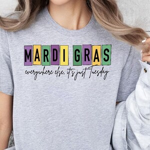May include: A light gray t-shirt with the text "Mardi Gras everywhere else, it's just Tuesday" in a colorful block design. The shirt is a short-sleeved crew neck.