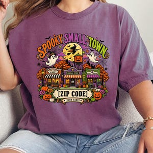 May include: A purple short-sleeve t-shirt featuring a colorful graphic of a small town with Halloween elements, including ghosts, a witch on a broomstick, pumpkins, and a water tower. The text reads SPOOKY SMALL TOWN, ZIP CODE, and TOWN NAME.