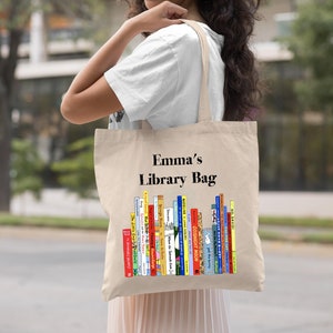 May include: A beige canvas tote bag with the text "Emma's Library Bag" printed in black. The bag features a colorful illustration of a stack of children's books.