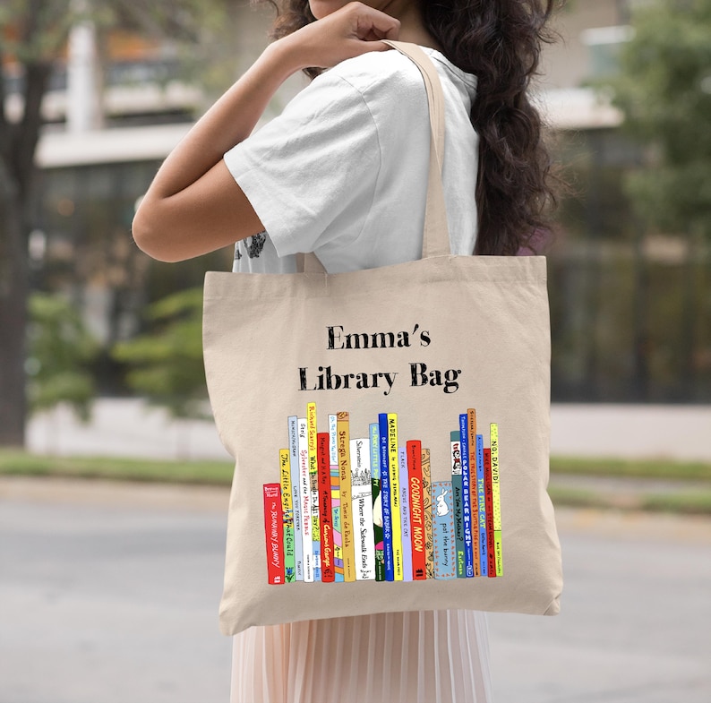 May include: A beige canvas tote bag with the text "Emma's Library Bag" printed in black. The bag features a colorful illustration of a stack of children's books.