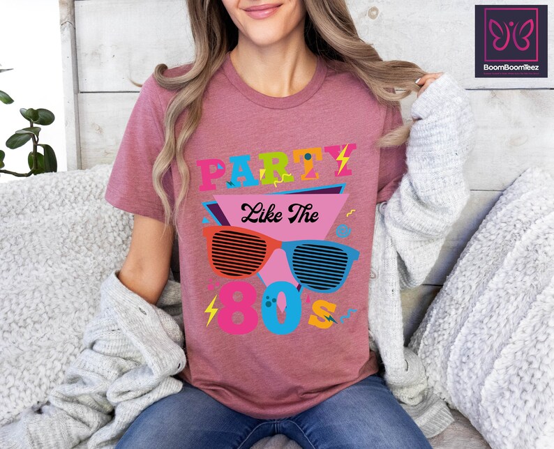 May include: A pink t-shirt with the text "Party Like The 80's" in colourful, retro-style lettering and a graphic of sunglasses with a lightning bolt design.