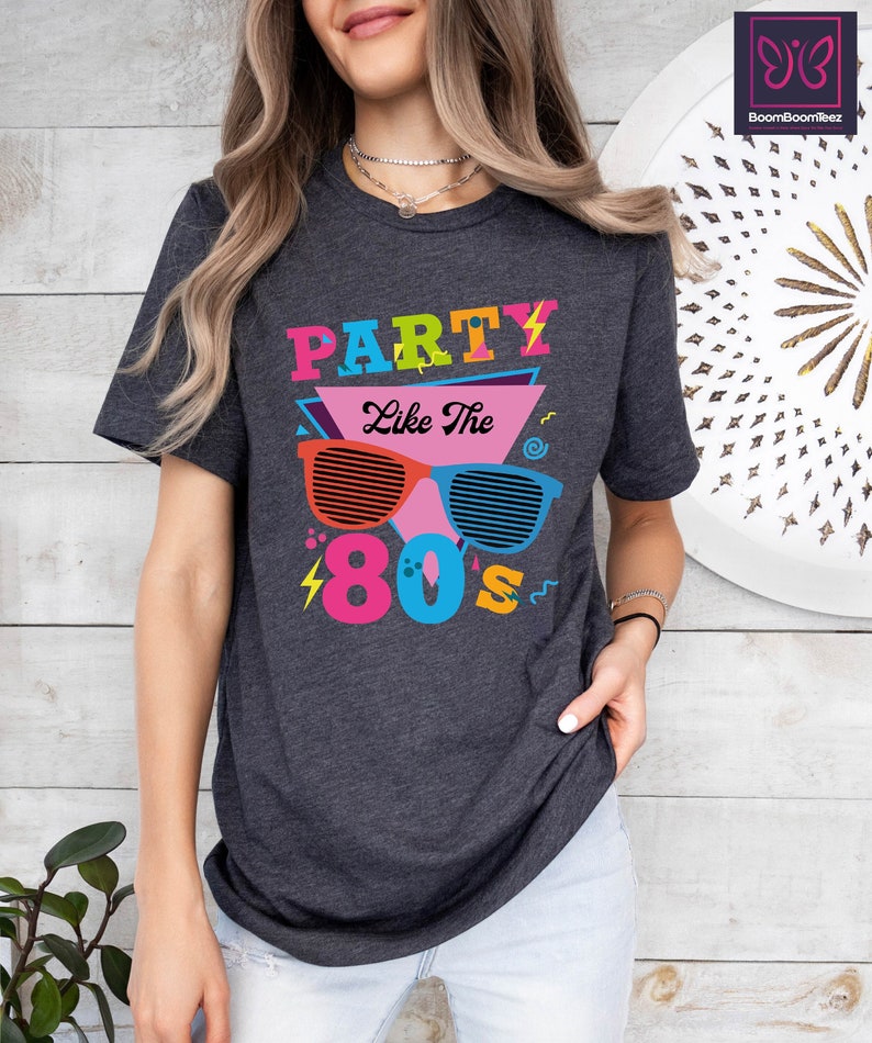May include: A dark grey t-shirt with a colourful graphic design that says "Party Like The 80's". The graphic features a pair of sunglasses with a lightning bolt and a musical note.