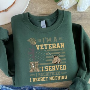 以下が含まれることがあります： 暗い緑色のスウェットシャツに、「I'm a Veteran I Served I Sacrificed I Regret Nothing」という文字が白と茶色の文字で書かれています。デザインには、軍用ヘルメット、十字架、ドッグタグ、アメリカ国旗も含まれています。