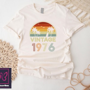 May include: Cream-colored t-shirt featuring a retro sunset design with palm trees, birds, and the text "VINTAGE 1976." The design incorporates shades of orange, yellow, green, and blue. The shirt has short sleeves and a crew neck.