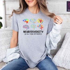 K&ouml;nnte beinhalten: Graues T-Shirt mit einem bunten Gehirndesign und dem Text "Neurodiversity We all think differently".