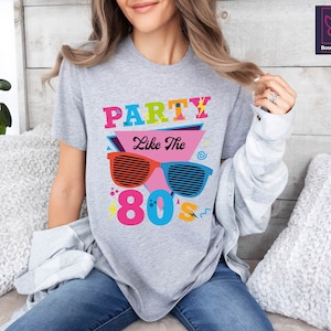 May include: A grey t-shirt with the text "Party Like The 80s" in colourful letters and a graphic of retro sunglasses.