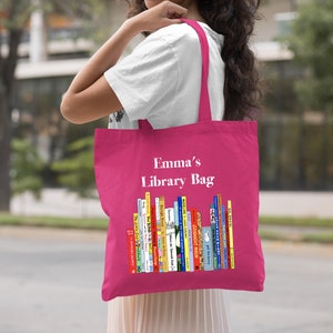 May include: A pink tote bag with the text "Emma's Library Bag" printed in white. The bag features a colorful illustration of a bookshelf filled with children's books.