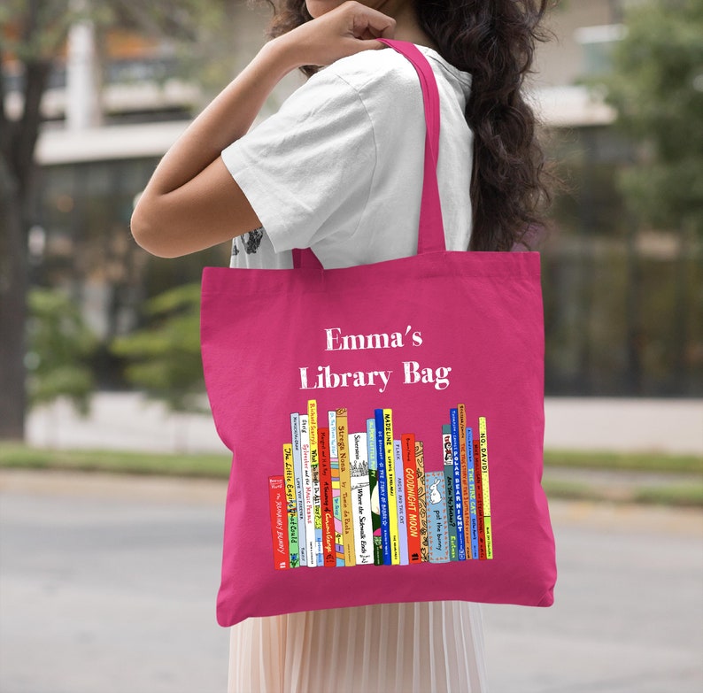 May include: A pink tote bag with the text "Emma's Library Bag" printed in white. The bag features a colorful illustration of a bookshelf filled with children's books.