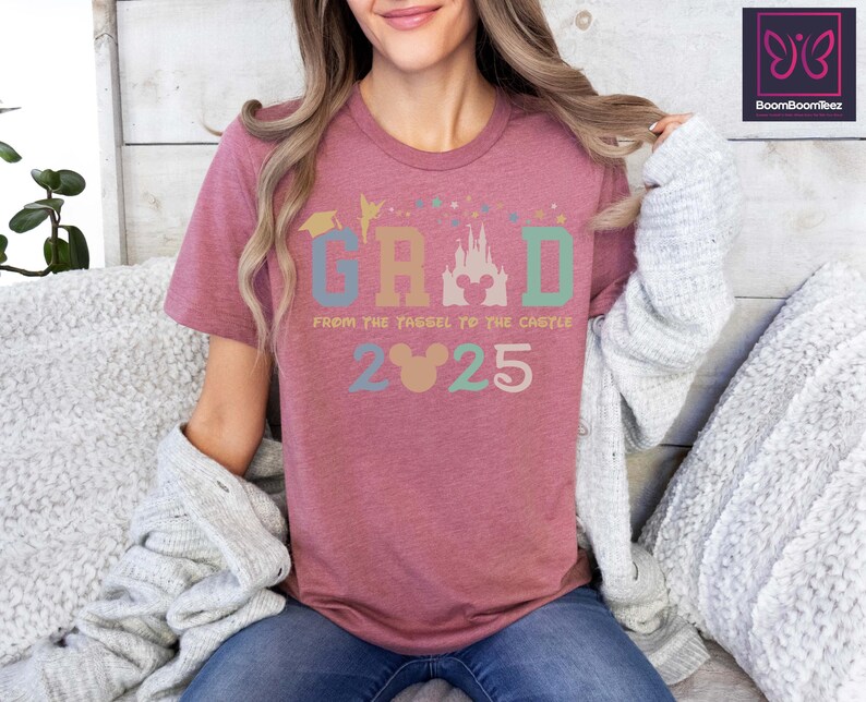 May include: A pink t-shirt with a colorful graphic that says "GRAD from the tassel to the castle 2025" with a Mickey Mouse head in the middle of the word "GRAD".