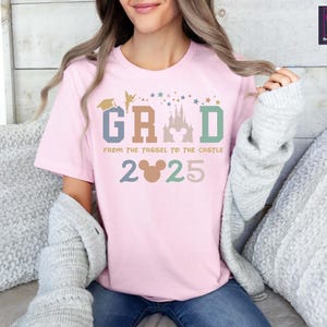 May include: Pink t-shirt with a graphic design that says "GRAD from the tassel to the castle 2025". The graphic includes a Mickey Mouse head, a graduation cap, Tinkerbell, and a silhouette of Cinderella's castle.