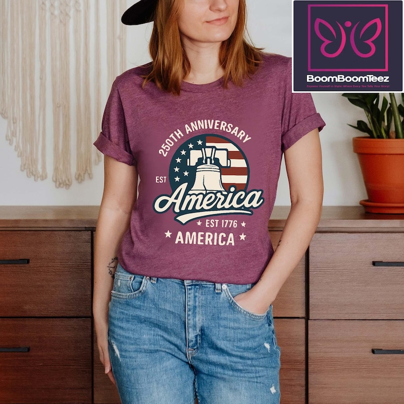 May include: Maroon t-shirt with a graphic celebrating the 250th anniversary of America. The design features the Liberty Bell, the American flag, and the text "America Est 1776". The shirt is paired with blue jeans.