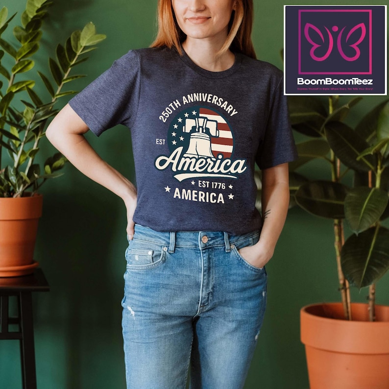May include: Navy blue t-shirt with a graphic commemorating the 250th anniversary of America. The design features the Liberty Bell, the American flag, and the word "America" in a vintage style. The shirt is worn with blue jeans.