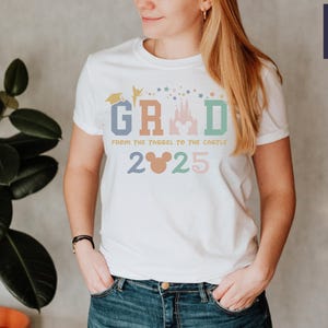 May include: White t-shirt with a colorful graphic that says "GRAD from the YAGEL to the CASTLE 2025". The graphic includes a graduation cap, a fairy, a castle, and Mickey Mouse ears.