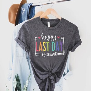May include: A dark grey t-shirt with the text "happy last day of school" in rainbow colours and white outlines. The text is framed with a white square and two small pink hearts.
