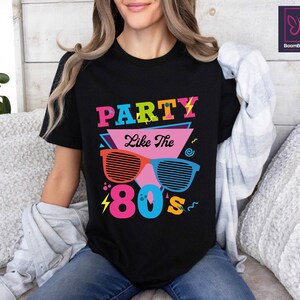 May include: Black t-shirt with a graphic design featuring the text "Party Like The 80's" in colourful letters and a pair of sunglasses with a retro design.