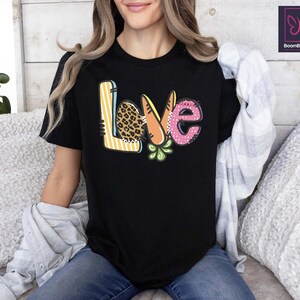 May include: Black t-shirt with the word "LOVE" in a colorful, patterned design. The "L" has a leopard print, the "O" is a carrot, the "V" is pink with polka dots, and the "E" has green leaves. The shirt is worn by a person.