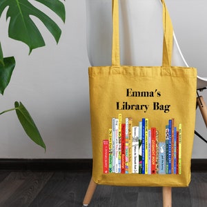 May include: A yellow canvas tote bag with the text "Emma's Library Bag" printed on the front. The bag features a colorful illustration of a bookshelf filled with books.