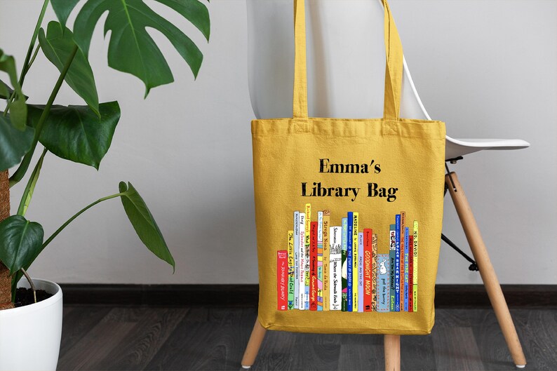 May include: A yellow canvas tote bag with the text "Emma's Library Bag" printed on the front. The bag features a colorful illustration of a bookshelf filled with books.