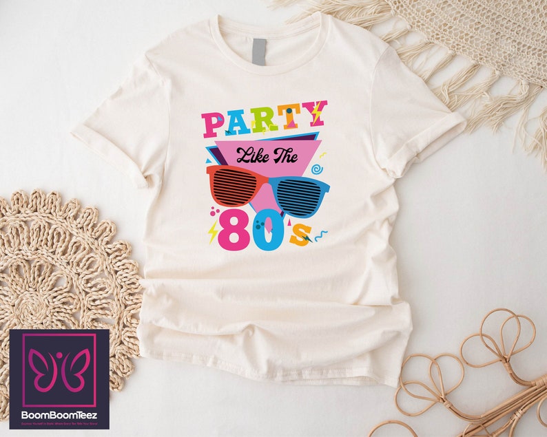 May include: A white t-shirt with a graphic design featuring the text "Party Like The 80s" in colourful letters and a pair of sunglasses with a retro design.