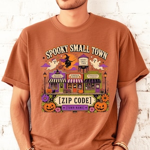 May include: A rust-colored t-shirt featuring a graphic print with the text SPOOKY SMALL TOWN, [ZIP CODE], and [TOWN NAME]. The design includes a witch on a broom, ghosts, a water tower, and three storefronts labeled Witch's Emporium, Pumpkin Bakery, and Curiosities.