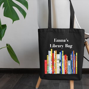 May include: Black canvas tote bag with white text that says "Emma's Library Bag". The bag has a colorful illustration of a bookshelf filled with books.