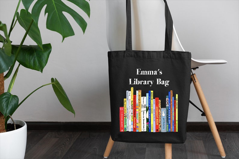 May include: Black canvas tote bag with white text that says "Emma's Library Bag". The bag has a colorful illustration of a bookshelf filled with books.