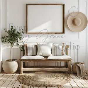 May include: A wooden bench with a white cushion and three pillows, two striped and one solid white. The bench is in front of a white wall with a framed picture and a straw hat hanging on the wall. A potted olive tree is on the left side of the bench. A woven rug is on the floor in front of the bench.