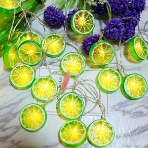 May include: A decorative string of lights with green and yellow lemon slice designs. The lights are strung on a thin, silver wire and emit a warm glow. Purple flowers and a green bottle are in the background. The text "ETERNITY BLOSSOM" is visible.