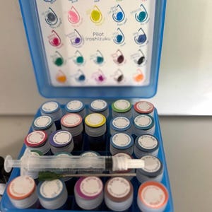 May include: A blue plastic storage case containing multiple small glass vials of Pilot Iroshizuku fountain pen ink. The lid features a color-coded chart of ink names. A clear plastic syringe with a metal needle rests horizontally across the vial caps.