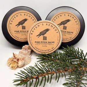Peut inclure: Trois boîtes de Pine Pitch Salve avec un logo de corbeau et du texte. Les étiquettes indiquent "Raven's Remedy" et "Lake County, BC". Un morceau de résine et une branche de pin sont également visibles.