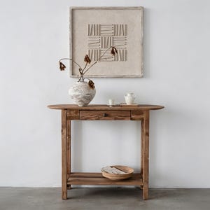 May include: A rustic wooden console table with an oval top, a drawer, and a lower shelf. A decorative vase with dried flowers sits on the table, along with a small white pitcher and cup. A framed artwork hangs on the white wall.