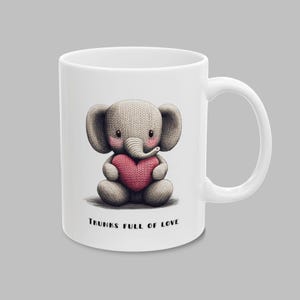 May include: White ceramic mug featuring a grey knitted elephant holding a pink heart. The mug has the text "Trunks Full of Love" printed on the bottom.