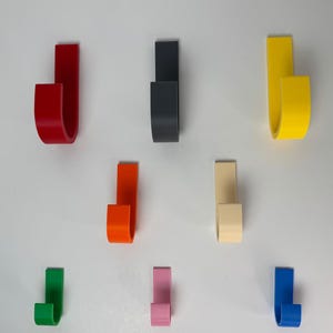 Könnte beinhalten: Neun farbige, J-förmige Haken in verschiedenen Farbtönen, darunter Rot, Schwarz, Gelb, Orange, Beige, Grün, Rosa und Blau. Die Haken sind vor einem weißen Hintergrund angeordnet und zeigen ihr gebogenes Design. Zum Aufhängen von Gegenständen.