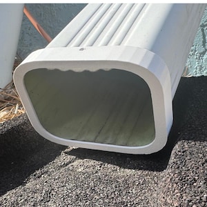 May include: A white rectangular gutter downspout with a rounded edge. The downspout is positioned over a black, textured surface. The interior of the downspout is a dark green colour.