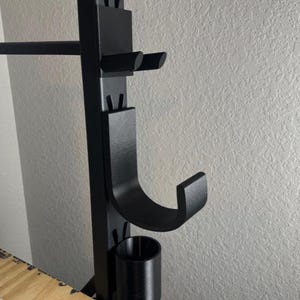 May include: Black weightlifting rack with multiple attachments. The rack has a bar holder, J-hooks, and a weight storage peg. The rack is made of metal and is mounted on a wall.