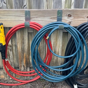 May include: A selection of garden hoses in red, blue, and black, suspended on a wooden fence. A yellow and black cordless drill is also mounted on the fence. The hoses are coiled, with metal connectors visible.