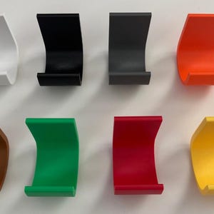 May include: A collection of eight curved, wall-mounted phone holders in various colors. The holders are white, black, gray, orange, brown, green, red, and yellow. Each holder has a curved design with a flat base.