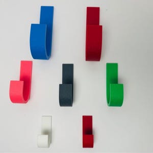 Puede incluir: Colección de coloridos ganchos de pared curvos en varios tonos, incluyendo azul, rojo, verde y blanco. Los ganchos están montados sobre una superficie blanca, mostrando su diseño simple y moderno. Estos ganchos son ideales para organizar objetos.