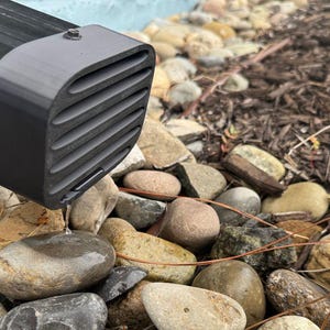 Könnte beinhalten: Ein schwarzes, rechteckiges Lüftungsgitter mit horizontalen Lamellen und einem abgerundeten Ende. Das Gitter befindet sich über einem Bett aus glatten, abgerundeten Steinen und braunem Mulch. Eine kleine Menge Wasser ist sichtbar, die aus dem Gitter tropft.