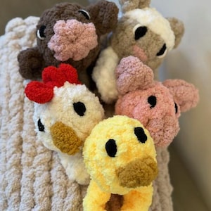 May include: Five crocheted stuffed animals in the shapes of a cow, pig, chicken, duck, and a brown and white animal. The animals are sitting on a beige knitted blanket.