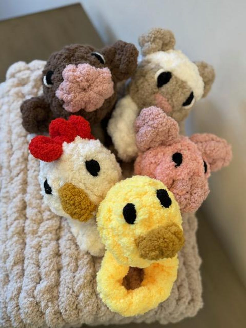 May include: Five crocheted stuffed animals in the shapes of a cow, pig, chicken, duck, and a brown and white animal. The animals are sitting on a beige knitted blanket.