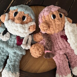 Op de afbeelding: Twee gehaakte pluche speelgoedjes, een in lichtblauw met een wit gezichtsmasker en de andere in roze met een wit gezichtsmasker. Beide speelgoedjes hebben bruine ogen en rode harten op hun borst.
