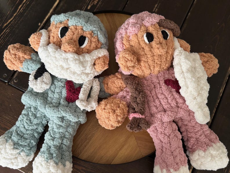 Op de afbeelding: Twee gehaakte pluche speelgoedjes, een in lichtblauw met een wit gezichtsmasker en de andere in roze met een wit gezichtsmasker. Beide speelgoedjes hebben bruine ogen en rode harten op hun borst.