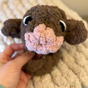 May include: A brown and pink crocheted stuffed animal with black eyes. The animal has a small, pink nose and is sitting on a white blanket.