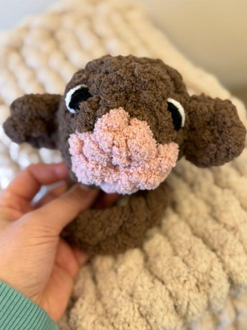 May include: A brown and pink crocheted stuffed animal with black eyes. The animal has a small, pink nose and is sitting on a white blanket.